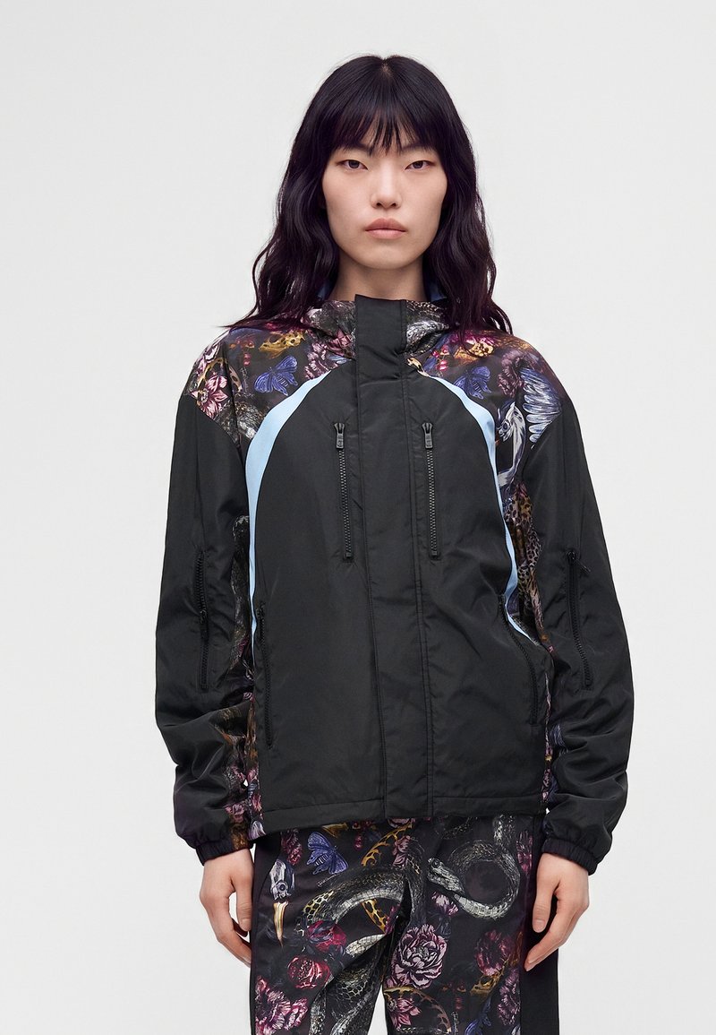 Femme aux longs cheveux noirs porte une veste noire avec des motifs floraux et papillons et un pantalon assorti avec des serpents et des fleurs.