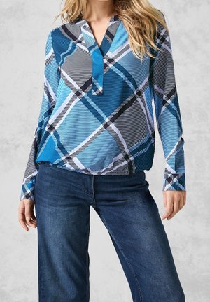 Kobieta ubrana w niebiesko-czarno-białą kraciastą bluzkę z długim rękawem i mankietami na guziki oraz ciemnoniebieskie dżinsy, stojąca na neutralnym tle.