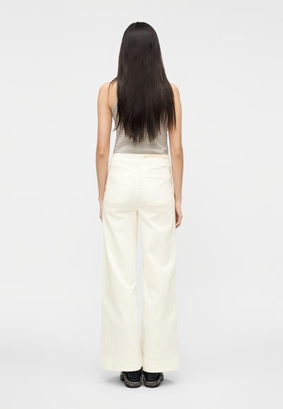 Pantalon blanc ample à taille haute et jambes larges, fabriqué en tissu doux. Associé à un débardeur rayé et des chaussures foncées.