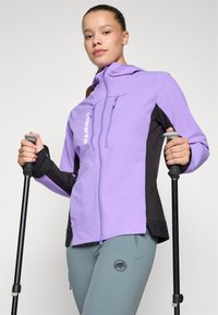 Violetinė ir juoda zip-up striukė su aukštu apykakle ir priekinėmis kišenėmis. Teksto logotipas ant krūtinės. Modelis laiko žygio lazdas, dėvi šviesiai mėlynas kelnes.