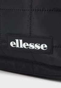 Czarna torba z podszewką i teksturowaną powierzchnią, z białym logo "ellesse" na czarnej łatce. Minimalistyczny design z pikowanymi sekcjami.