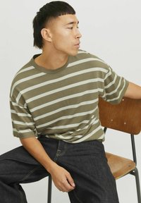 Olivengrøn stribet t-shirt med hvide striber, rund halsudskæring, korte ærmer, parret med mørke jeans. Siddende på en træstol.