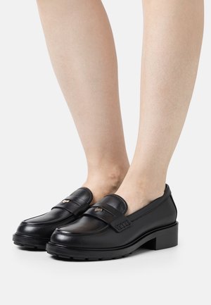 Mocasines de cuero negro con un bajo tacón cubano, que presentan una correa en la parte frontal y costuras decorativas. Textura suave y acentos sutiles.