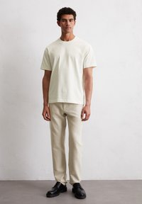 Light-colored cotton T-shirt with short sleeves, paired with beige trousers and black lace-up shoes on a neutral background.