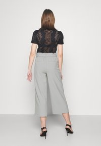 Kvinna i svart spetstopp med korta ärmar, ljusgrå hög midja capribyxor och svarta högklackade sandaler står med ryggen mot kameran.