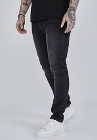 Black slim-fit jeans made from denim, featuring a smooth texture with slight fading. Paired with white sneakers and visible tattoos on the arm.