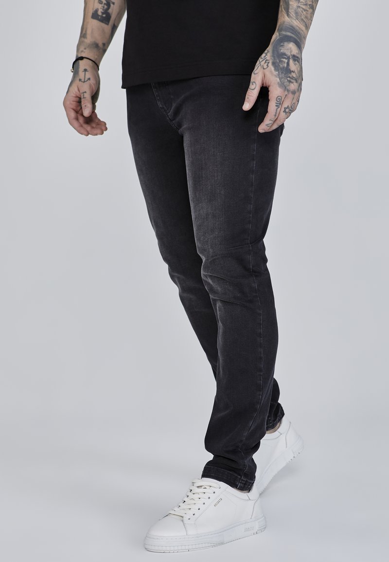 Black slim-fit jeans made from denim, featuring a smooth texture with slight fading. Paired with white sneakers and visible tattoos on the arm.