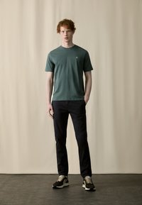 T-shirt en coton vert à manches courtes, col rond, avec un petit logo sur la poitrine, associé à un pantalon sombre et des baskets multicolores.
