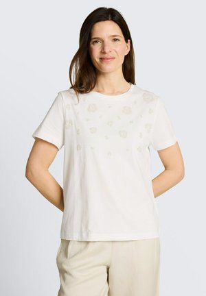 Vrouw met schouderlang bruin haar, die een wit shirt met korte mouwen draagt met subtiele bloemenpatronen en beige broek, glimlacht zacht.