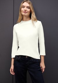Top blanco de manga larga con cuello alto, combinado con jeans de mezclilla oscuros. Tela suave con un corte limpio y moderno. Ajuste casual y diseño minimalista.