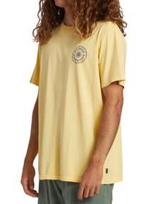 Camiseta de algodón amarilla con mangas cortas, que presenta un gráfico circular en gris en el pecho. Corte relajado, diseño simple y sin patrones visibles.