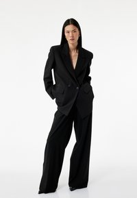 Sort skræddersyet blazer med store revers, dobbelte knapper og for-poser, parret med løse sorte bukser og spidse sorte hæle.