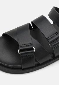 Svarta sandaler med tre breda syntetiska remmar med texturerad yta, fästa med ringar i silverfärgad metall, på en slät svart sula.