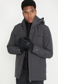 Grå isolerad jacka med huva, dragkedja framtill och knappdekorationer, som bärs över ett svart innerlager; modellen håller svarta handskar.