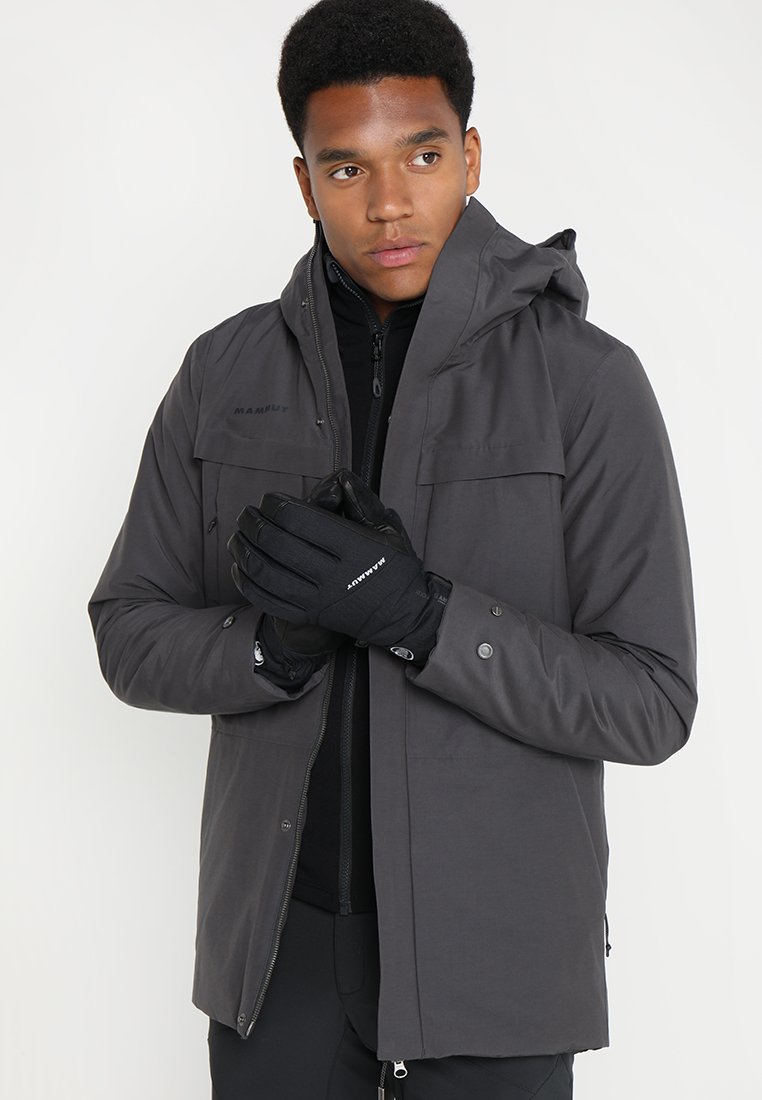 Grå isolerad jacka med huva, dragkedja framtill och knappdekorationer, som bärs över ett svart innerlager; modellen håller svarta handskar.