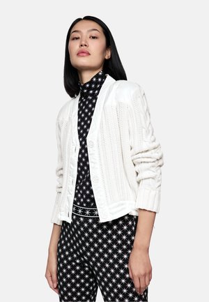 White knitted cardigan with cable patterns, featuring a satin shoulder and front buttons, paired with a black patterned top and pants.