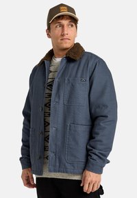 Blue, button-up jacket with a brown, fleece-lined collar, two front pockets, and a textured gray patterned sweater underneath.