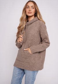 Brązowa, dzianinowa bluza z kapturem z prążkowanym dołem i mankietami, kieszenią kangurzą oraz sznurkiem przy szyi. Miękka tekstura, luźny krój, w zestawieniu z dżinsem.