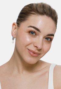 Silberne Ohrringe mit einem Kettenlink-Design und einem Perlenanhänger. Die Person hat helle Haut, schulterlanges, nach hinten gekämmt Haar und blaue Augen.