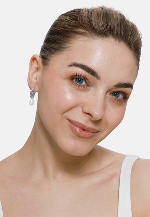 Silberne Ohrringe mit einem Kettenlink-Design und einem Perlenanhänger. Die Person hat helle Haut, schulterlanges, nach hinten gekämmt Haar und blaue Augen.