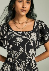 Vestito nero floreale con motivi botanici bianchi, maniche a volant e bordi in pizzo. La modella indossa collane d'oro a strati e orecchini a cerchio.