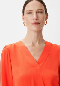Leuchtend orange Bluse mit V-Ausschnitt und langen Ärmeln. Glattes, strukturiertes Gewebe mit subtiler glänzender Oberfläche, das an den Schultern geraffte Details aufweist.