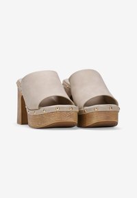 Mules de cuero beige destalonados con un chunky tacón de madera, que cuentan con adornos de tachuelas doradas a lo largo del costado y una plataforma de madera texturizada.