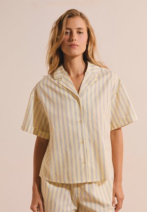 Femme aux cheveux blonds lâchés portant une chemise jaune pâle à manches courtes et un pantalon à rayures verticales bleues et blanches.