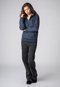 Marine blauwe ritshoodie met een gestructureerde afwerking en voorzakken, gecombineerd met zwarte jeans en zwarte schoenen. De capuchon heeft koorden.