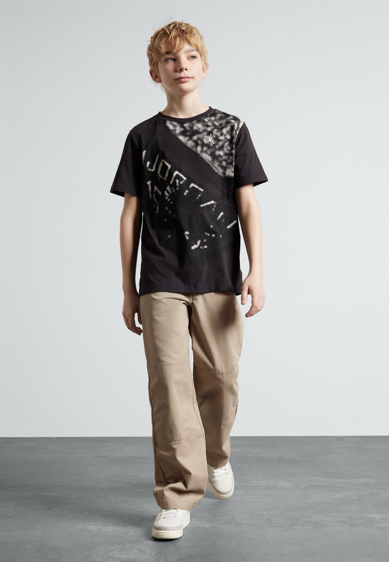 T-shirt nera a maniche corte con un motivo testurizzato su un lato, abbinata a pantaloni cargo color sabbia e sneakers bianche. Taglio semplice e rilassato.