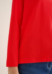 Top rosso a maniche lunghe realizzato in un tessuto leggero, con una texture liscia e un taglio dritto, abbinato a jeans di denim.