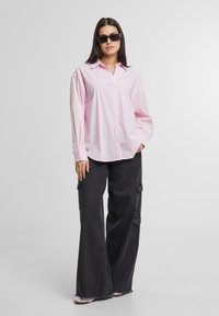 Roze en witte gestreepte button-up shirt met een relaxte pasvorm, gecombineerd met wijdvallende zwarte cargobroek en donkere zonnebril. Glad textuur, casual uitstraling.