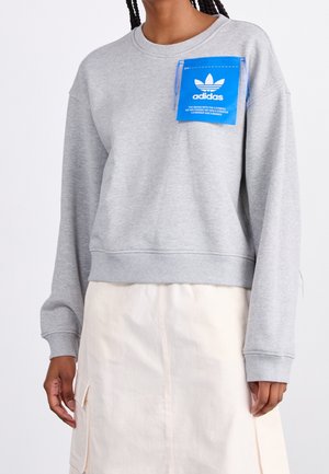 Osoba ubrana w jasnoszarą bluzę Adidas z niebieską naszywką z logo na piersi i kremową spódnicę z kieszeniami po bokach.