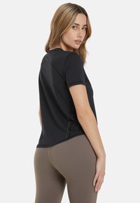 Črna atletska majica s kratkimi rokavi z okroglo izreza, brezšivna zasnova in majhne okrasne podrobnosti blizu roba. Lahek, prilegajoč material.