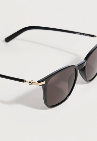 Μαύρα γυαλιά ηλίου με τετράγωνο πλαίσιο, σκουρόχρωμους φακούς και χρυσές λεπτομέρειες στις μεντεσέδες, σε λευκό φόντο.