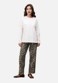 Vrouw die staat in een witte langetermijnblouse, met een luipaardprint broek en bruine platte schoenen voor een effen lichte achtergrond.