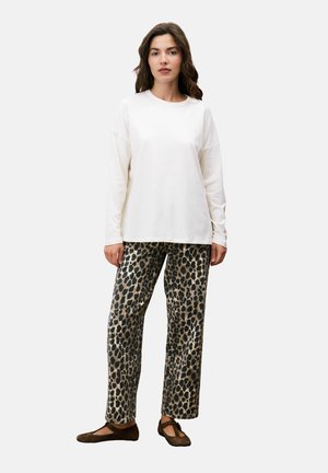 Vrouw die staat in een witte langetermijnblouse, met een luipaardprint broek en bruine platte schoenen voor een effen lichte achtergrond.