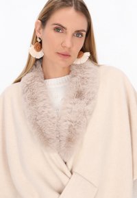 Palton bej cu guler din blană sintetică, textură moale și un pulover tricotat confortabil dedesubt. Cercei statement cu un design franjurat.