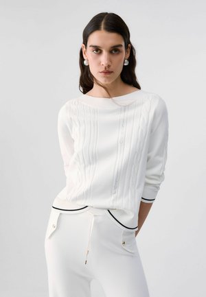 Donna con capelli lunghi e scuri che indossa un maglione bianco strutturato con bordi neri e pantaloni bianchi con tasche con bottoni, in piedi davanti a uno sfondo semplice.