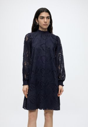 Γυναίκα με μακριά σκούρα μαλλιά που φοράει ένα μπλε δαντελένιο φόρεμα με μακριά μανίκια και ψηλό λαιμό, στέκεται μπροστά σε ένα απλό ανοιχτό φόντο.