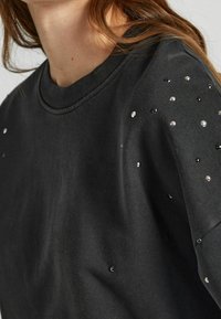 Mujer vistiendo una camisa negra de manga larga adornada con pequeñas cuentas plateadas y negras en el hombro y la parte superior de la manga.