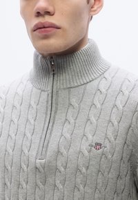Γκρι πουλόβερ με πλεκτό σχέδιο καλώδιο, ψηλό ριμπ κολάρο, μισό φερμουάρ και μικρό κέντημα με σχέδιο ασπίδας στο στήθος.