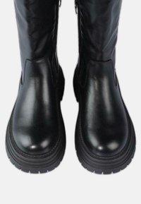 Par de botas de cuero negro brillante con suelas gruesas y con dibujo, erguidas y con cremalleras laterales visibles, sobre un fondo blanco.