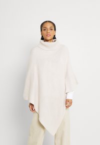 Poncho beige chiaro con un alto collo a coste. Il morbido tessuto a maglia testurizzato cade in modo elegante, assottigliandosi fino a un orlo appuntito nella parte anteriore.
