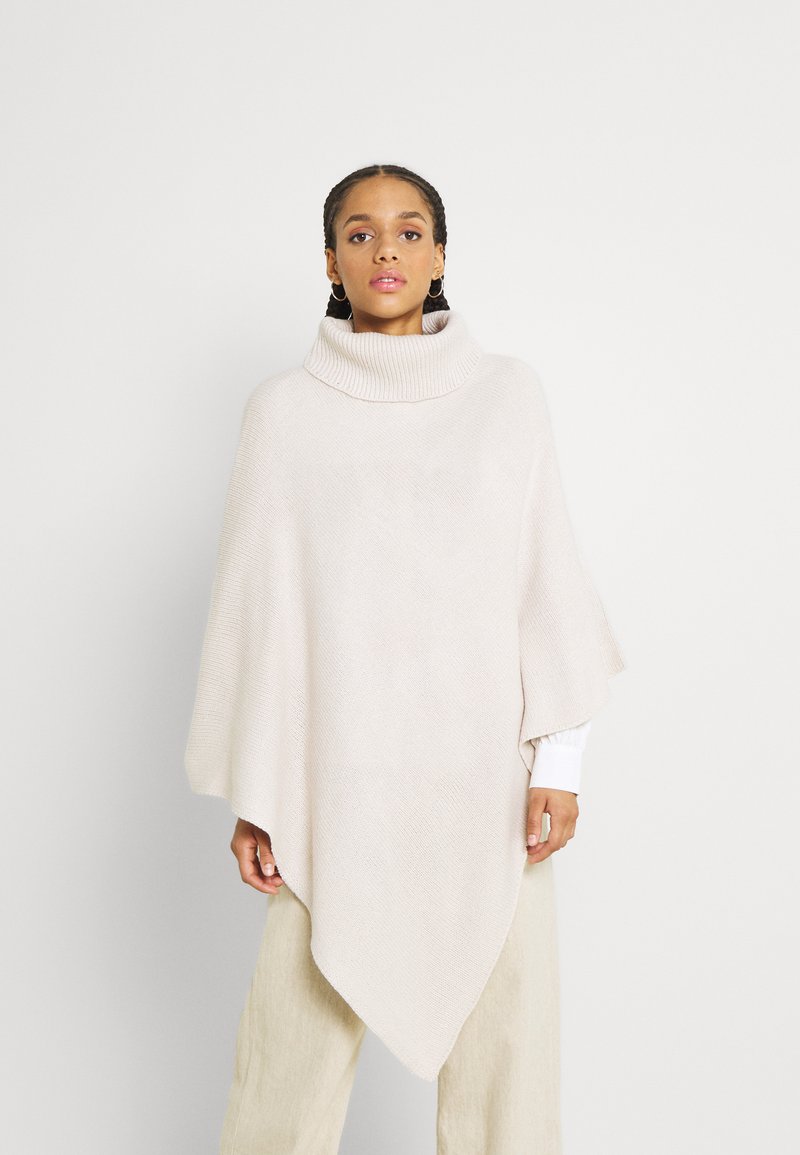 Poncho beige chiaro con un alto collo a coste. Il morbido tessuto a maglia testurizzato cade in modo elegante, assottigliandosi fino a un orlo appuntito nella parte anteriore.