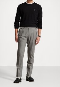 Suéter negro de manga larga, pantalones grises con pliegues y mocasines negros. El suéter tiene un escote redondo y un logo sutil; los pantalones presentan un ajuste a medida.