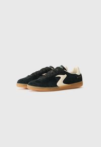 Black suede sneakers with a cream side swoosh, textured surface, black laces, and a gum rubber sole. Minimal branding on the heel.