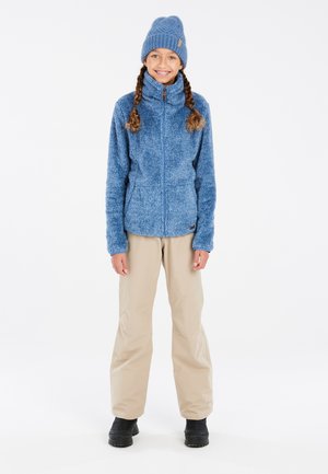 Blauwe fleecejas met hoge kraag, ritssluiting en gestructureerd oppervlak. Gecombineerd met beige broeken en zwarte waterdichte laarzen.