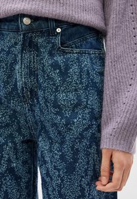 Κοντινό ενός ατόμου που φοράει μπλε τζιν με σχέδια και ένα μωβ πλεκτό πουλόβερ, με εμφανές χέρι που ακουμπάει στο πλάι.