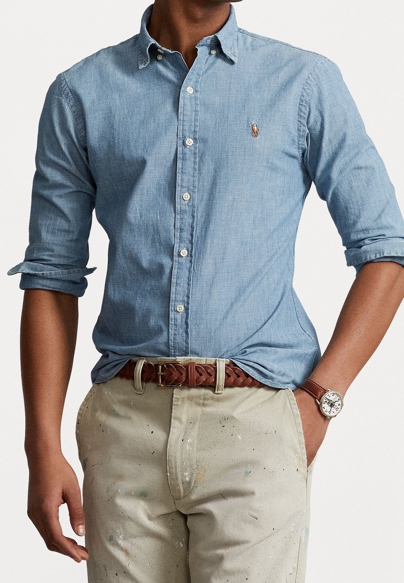 Camicia di jeans azzurro chiaro con bottoni e maniche arrotolate, con logo sul petto. Indossata con pantaloni beige macchiati di vernice e una cintura marrone.